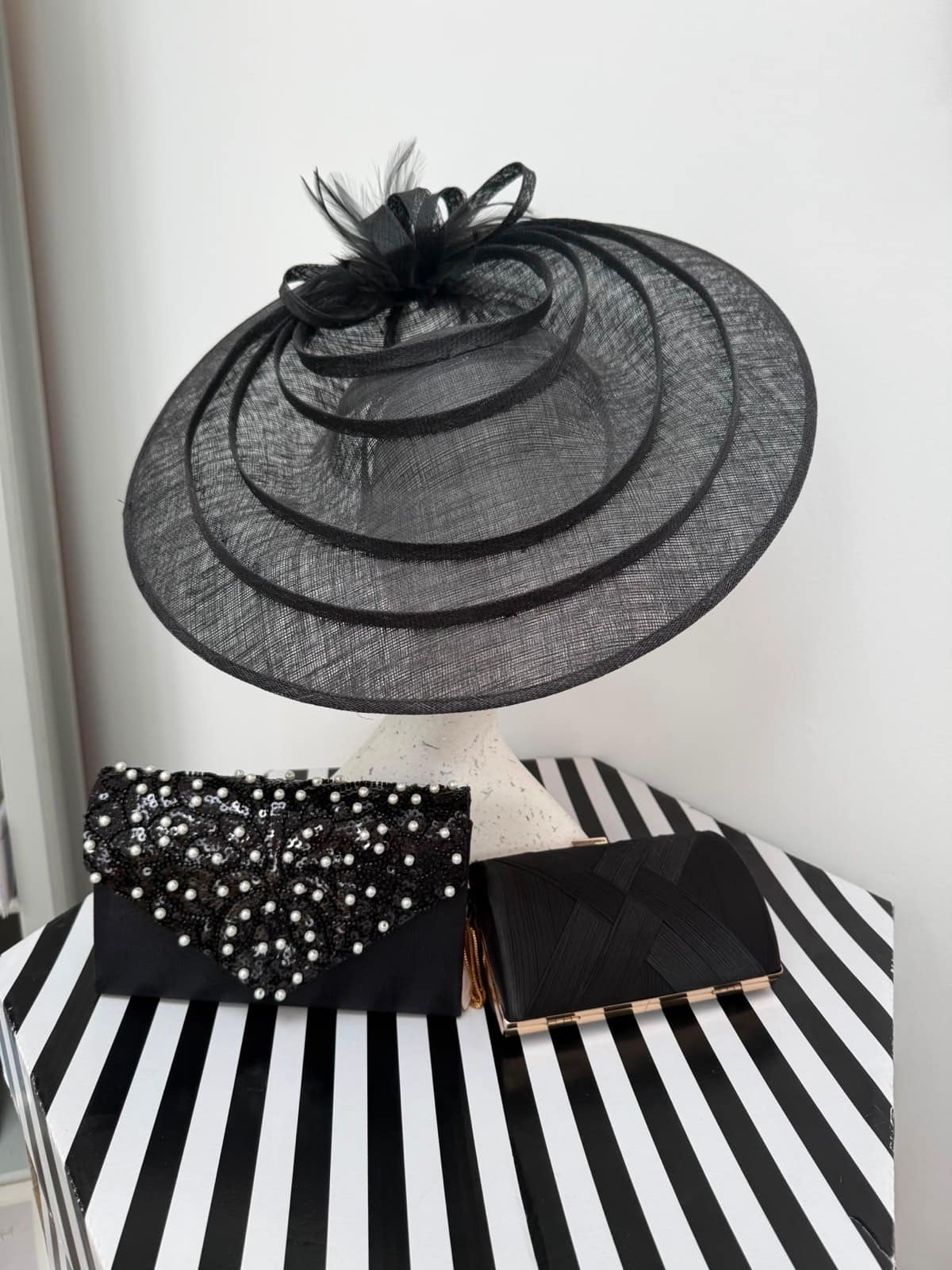 Black tiered sinamay saucer hat with concentric brims and feather detail, with black clutch bags