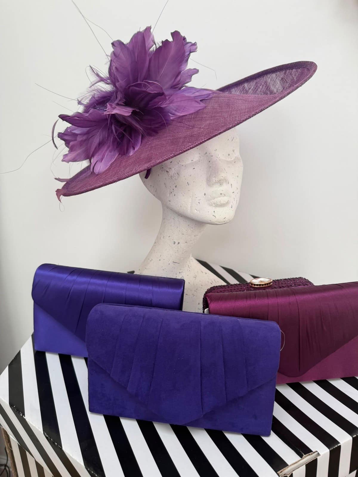 Amethyst wide-brim hatinator with a full feather flower, shown with purple and plum clutch bags