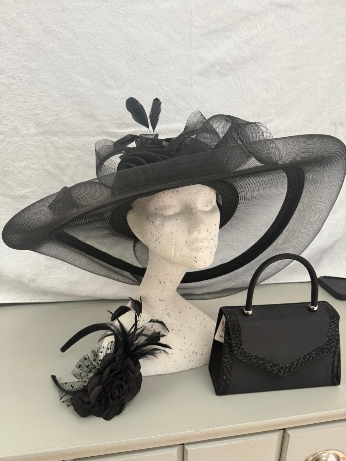 Black sheer-brim occasion hat with rose detail, black rose fascinator and black handbag