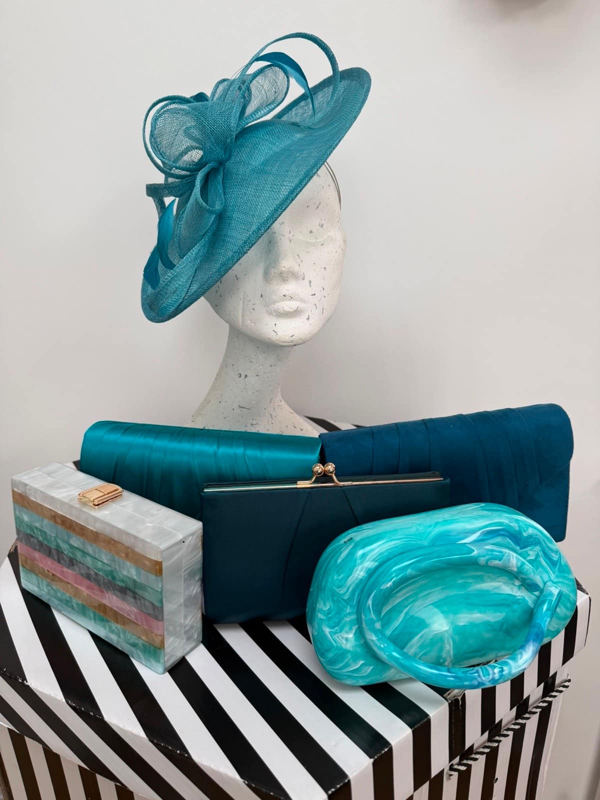 Turquoise saucer hatinator with looping bows, displayed with teal clutches and a marbled aqua handbag