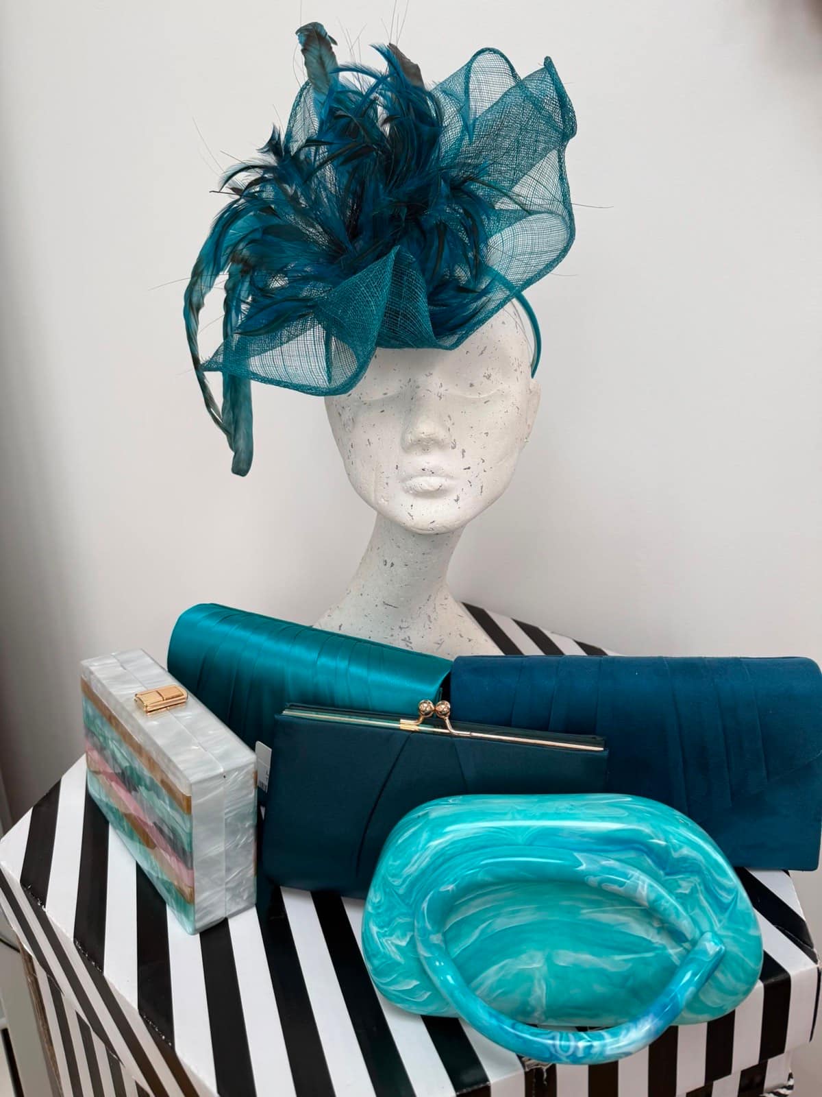 Deep teal crin and feather fascinator on a mannequin with teal satin and suede clutches